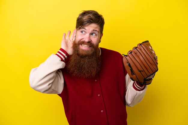 Homme joueur rousse avec barbe avec gant de baseball isolé sur fond jaune écoutant quelque chose en mettant la main sur l'oreille