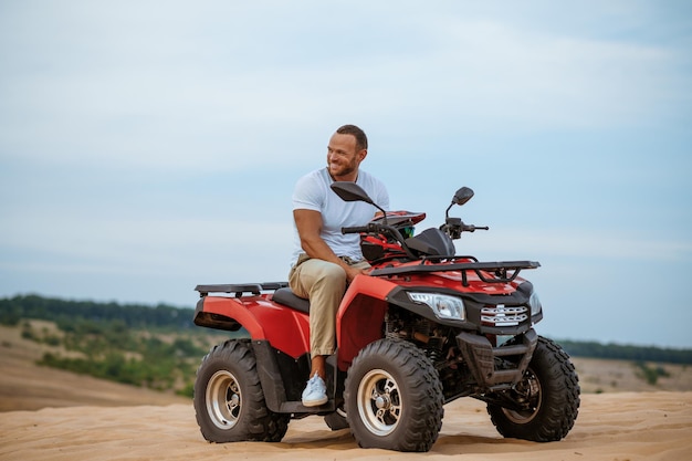 Un homme joyeux avec un casque à la main pose sur un vtt, une descente dans les sables du désert. Personne de sexe masculin en quad, course de sable, safari dans les dunes par une chaude journée ensoleillée, aventure extrême en 4x4, quad