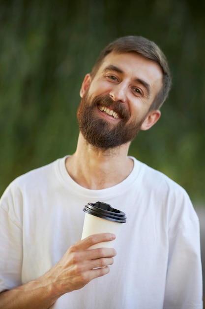 Un homme joyeux dans un T-shirt blanc boit du café dans une tasse en papier blanc. Un gobelet en papier blanc