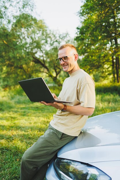 Un homme joyeux à lunettes est assis sur le capot de sa voiture dans la nature et travaille sur un ordinateur portable Travail indépendant travail à distance pendant les vacances