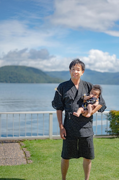 Un homme en kimono tient un bébé devant un lac.