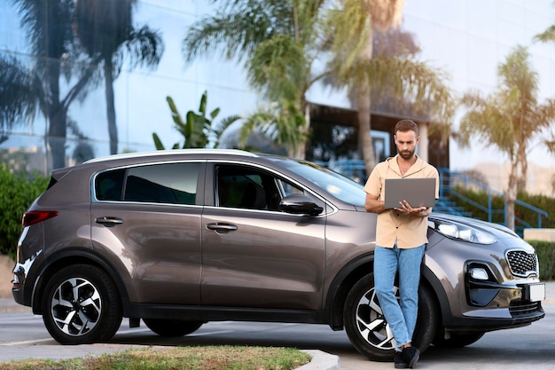 Homme latin d'âge moyen pensif utilisant un ordinateur portable, travaillant en ligne debout près d'une nouvelle voiture