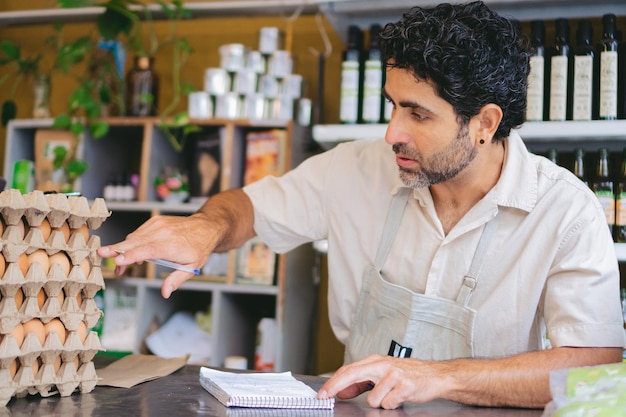 Photo homme latin d'âge moyen propriétaire d'une épicerie biologique vendant des œufs faisant de la comptabilité sur un papier