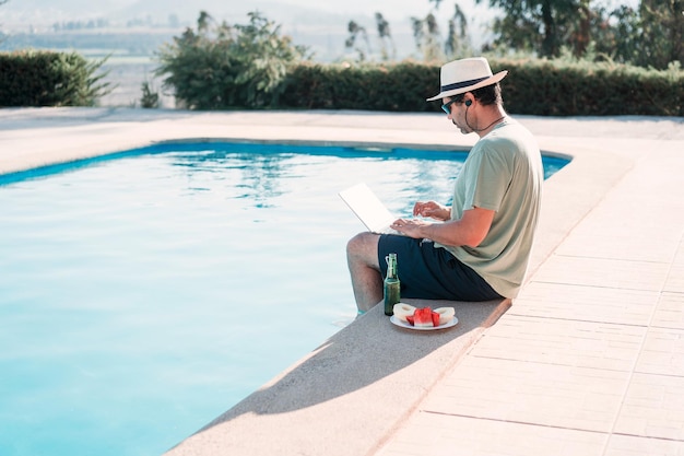 Homme latin assis au bord de la piscine avec un ordinateur portable et de la bière seul