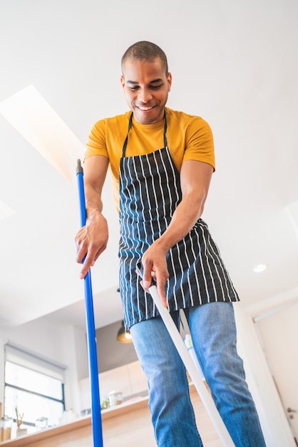 Homme latin balayant le plancher en bois avec balai à la maison.