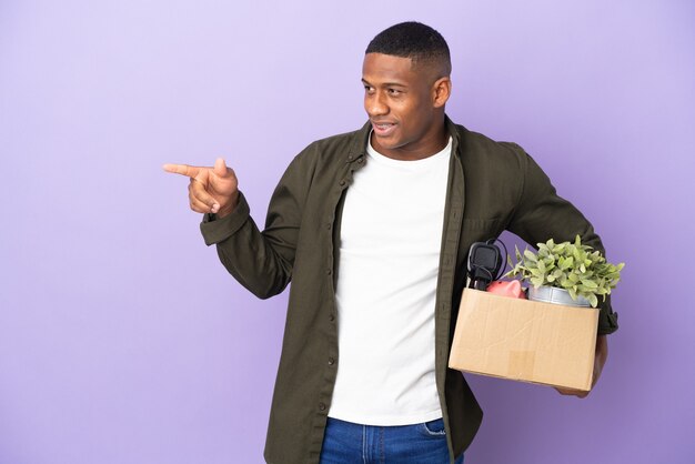 Homme latin faisant un mouvement tout en ramassant une boîte pleine de choses pointant le doigt sur le côté et présentant un produit