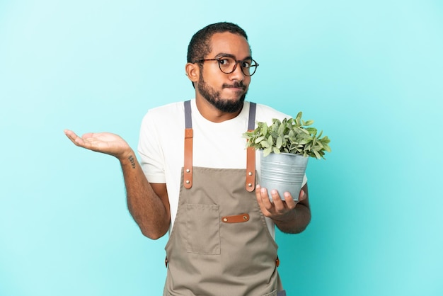 Homme latin jardinier tenant une plante isolée sur fond bleu ayant des doutes tout en levant les mains