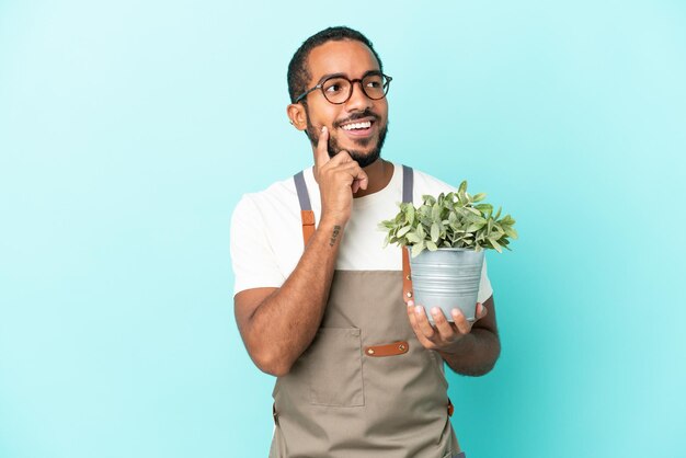 Homme latin jardinier tenant une plante isolée sur fond bleu pensant à une idée tout en levant les yeux