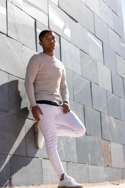 Homme latin sérieux posant dans un pantalon et un pull blanc, fond dallé vertical