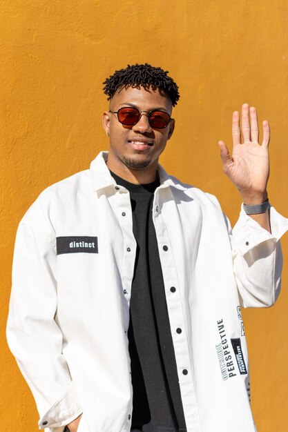 homme latin souriant avec des lunettes de soleil en agitant la main dans un fond jaune