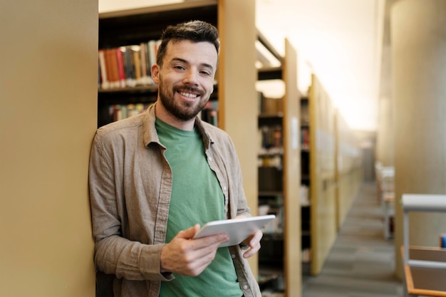 Homme latin souriant tenant une tablette numérique regardant la caméra Étudiant hispanique étudiant à la bibliothèque