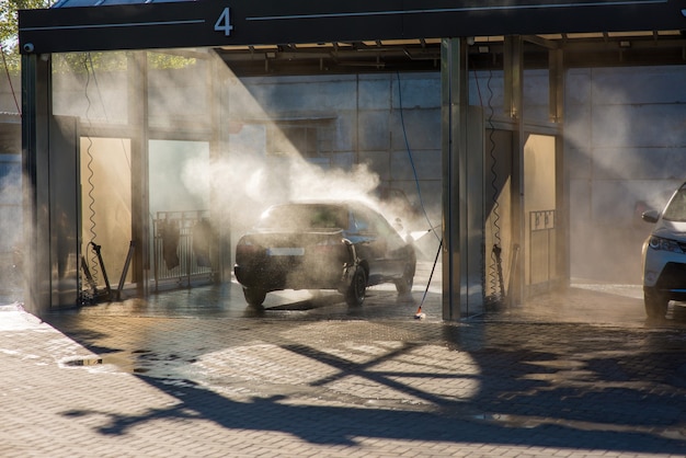 Un homme lave sa voiture avec un jet d'eau dans un lave-auto en libre-service dans les rayons arrière du soleil