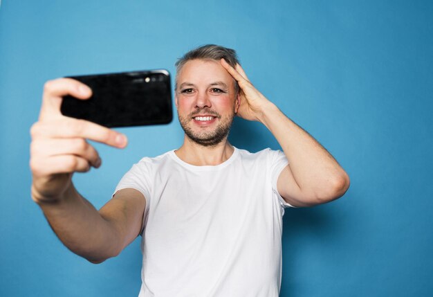 Un homme LGBT sourit et prend des photos de lui-même sur un appareil photo de smartphone debout sur un fond bleu isolé