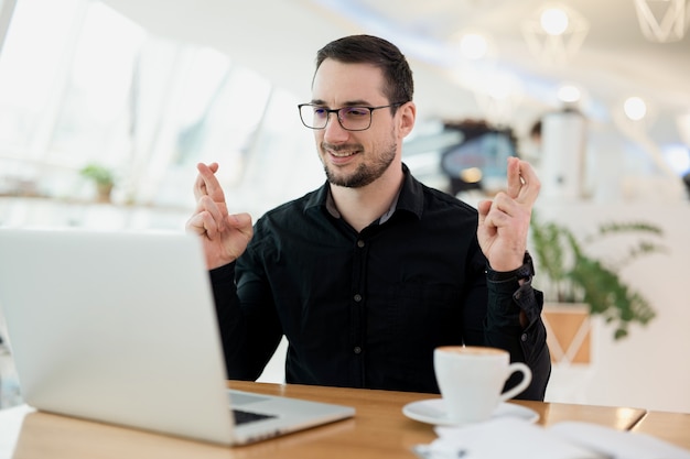 Homme à lunettes et chemise noire gesticulant les doigts croisés et souriant. Chance et concept superstitieux. Jeune homme séduisant avec barbe assis au café, parlant par caméra sur son ordinateur portable.