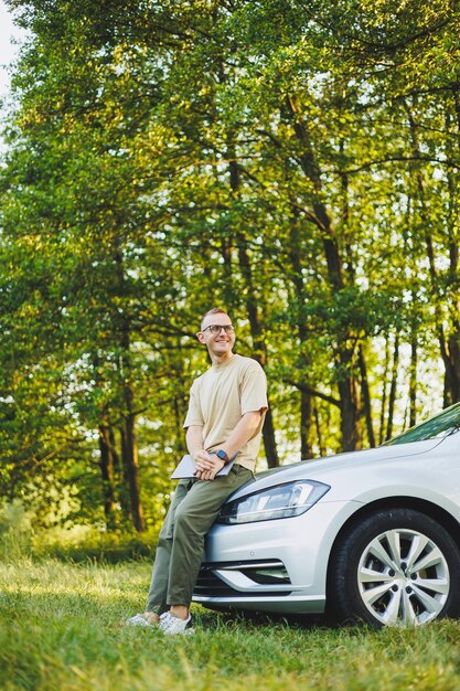 Un homme à lunettes qui réussit travaille sur un ordinateur portable tout en s'appuyant sur le capot de sa voiture. Travail à distance dans la nature Travailler sur un ordinateur portable en ligne Travailler sur un ordinateur en voyage