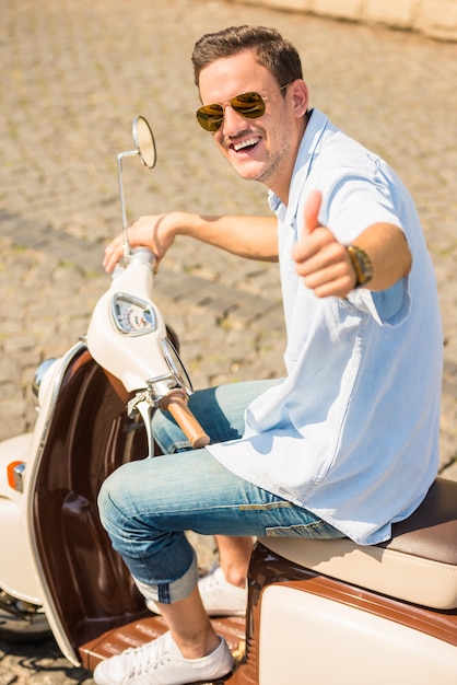 Homme à lunettes de soleil est assis sur un scooter et souriant.