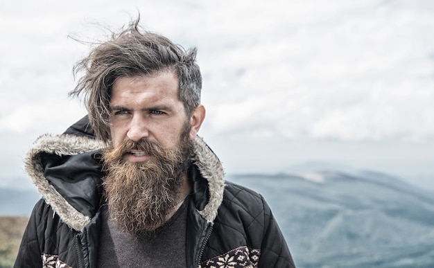 Homme macho barbu brutal. Bel homme hipster ou mec avec barbe et moustache sur un visage sérieux en chapeau et veste en plein air au sommet de la montagne contre ciel nuageux sur fond naturel