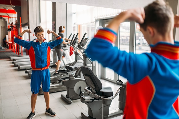 Homme maigre en vêtements de sport regarde ses muscles dans un miroir à la salle de sport, humour. Personne de sexe masculin faible dans le club de sport