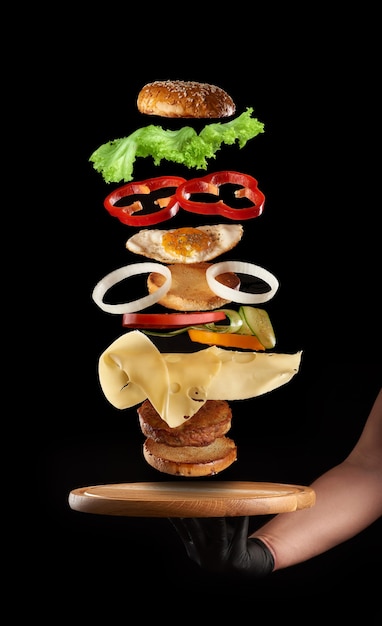 Homme main tenant une planche de bois ronde et des couches flottantes de cheeseburger avec oeuf de poulet et escalope de viande sur fond noir, restauration rapide