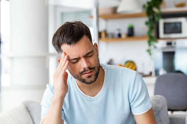 Homme à la maison ayant mal à la tête Jeune homme souffrant de maux de tête après une dure journée de travail assis sur un canapé à la maison