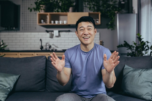 Homme à la maison regardant une webcam et souriant saluant des amis conférence en ligne Asiatique à la maison assis sur un canapé parlant à distance