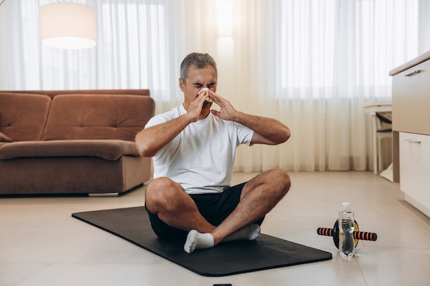 Homme malade âgé avec le nez qui coule assis sur un tapis de yoga noir