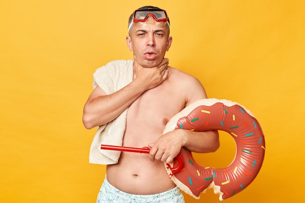 Homme malade avec des lunettes de plongée et un snorkel tenant un anneau en caoutchouc de donut isolé sur un fond jaune souffrant d'un mal de gorge se baigner trop longtemps dans de l'eau de mer froide