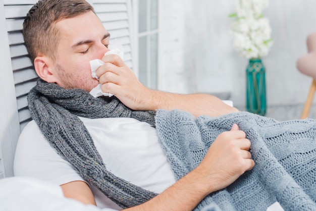 Homme malade se moucher avec du papier de soie blanc se trouvant sur le lit