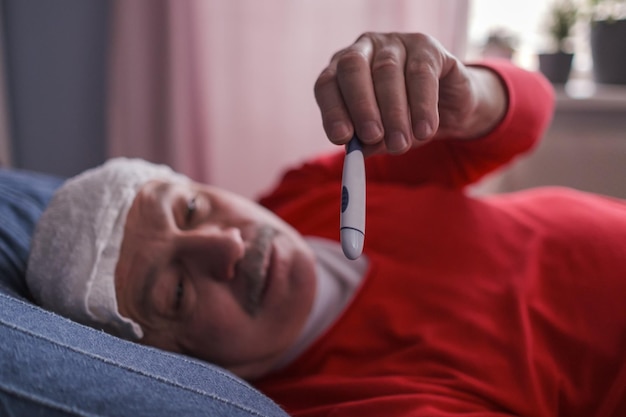 Un homme malade vérifiant le thermomètre allongé avec une serviette humide sur son front