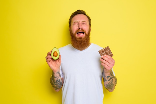 L'homme malheureux avec la barbe et les tatouages tient l'avocat et le chocolat