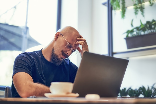 Un homme malheureux tient la tête dans la main, un homme d'affaires fatigué est assis au bureau avec un ordinateur portable qui a mal à la tête