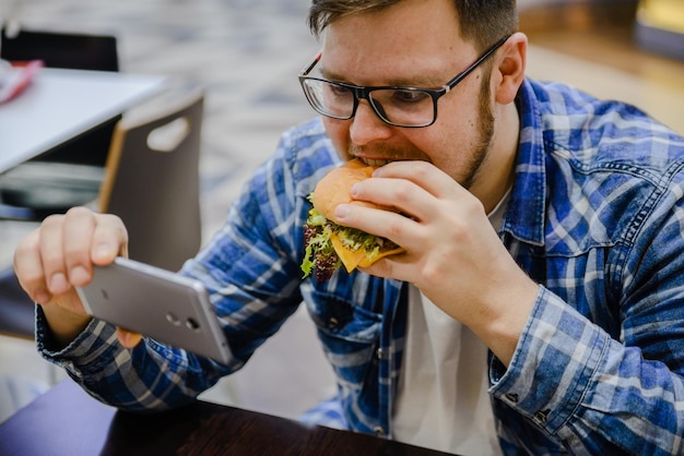 Un homme mange un hamburger au café en regardant une vidéo sur son téléphone