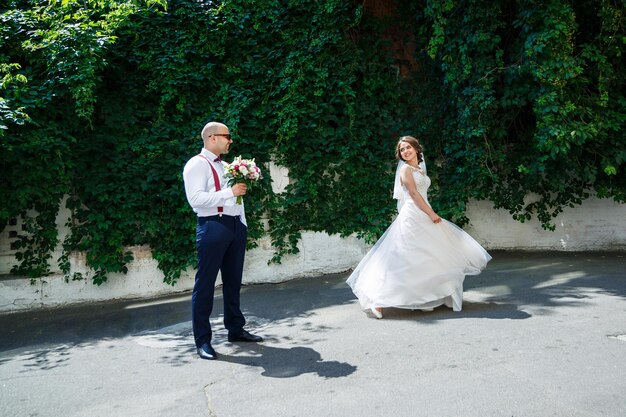 Homme marié dans une chemise et un pantalon blancs, une belle mariée dans une robe et un voile blancs, posant et marchant le long de la rue un jour de mariage