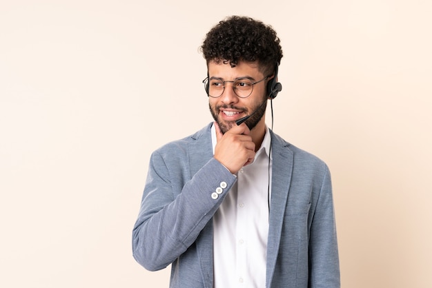 Homme marocain télévendeur travaillant avec un casque isolé sur beige à la recherche sur le côté et souriant