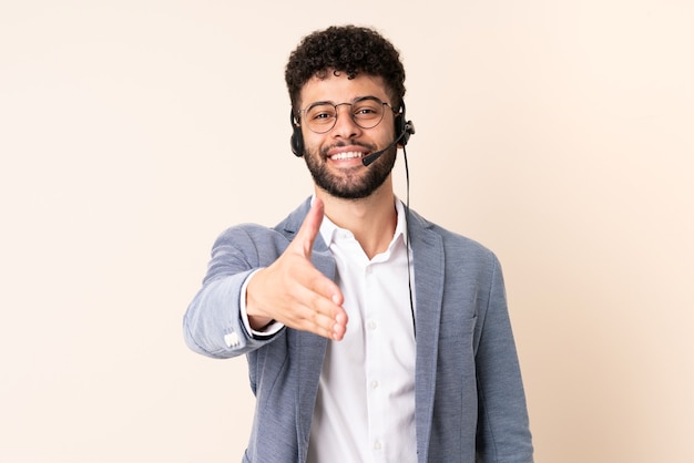 Homme marocain télévendeur travaillant avec un casque isolé sur mur beige se serrant la main pour conclure une bonne affaire