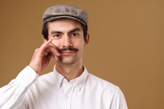 Photo homme masculin portant un chapeau regardant à l'avant et touchant sa moustache isolé sur beige