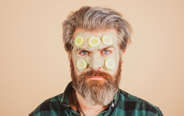 Homme avec un masque d'argile et des tranches de concombre sur le visage Spa dermatologie bien-être et concept de traitement du visage Homme ayant un masque hydratant cosmétique fond de studio isolé Soins du visage masculin