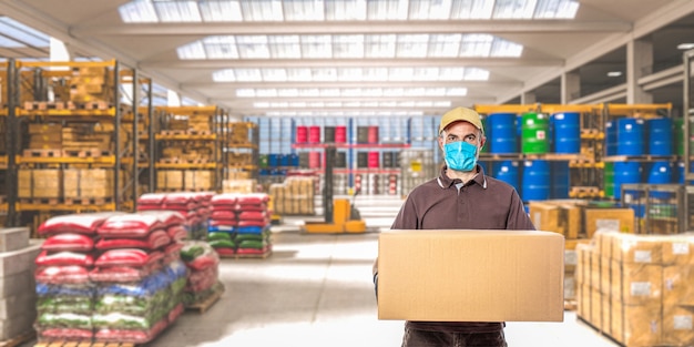 Homme avec masque et colis, intérieur d'un entrepôt industriel où différentes marchandises sont stockées.