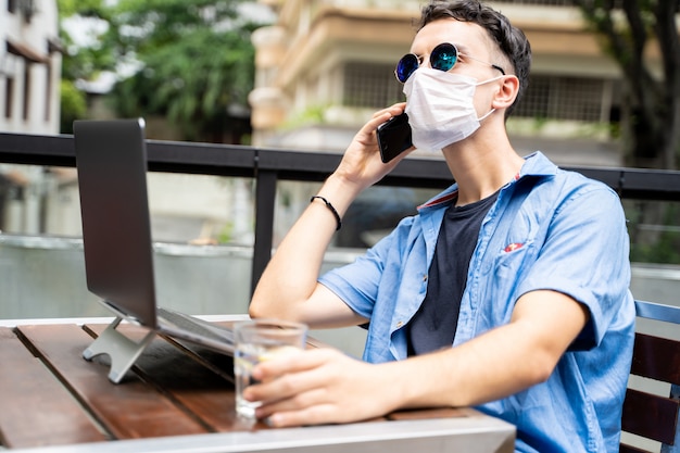 Homme avec masque et lunettes de soleil travaillant à distance avec son ordinateur portable et parlant à son téléphone portable à l'extérieur