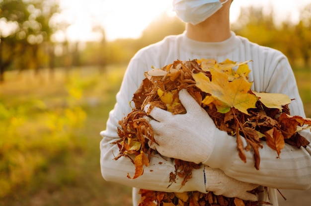 L'homme en masque médical de protection nettoie les feuilles d'automne dans le parc L'homme en gants recueille