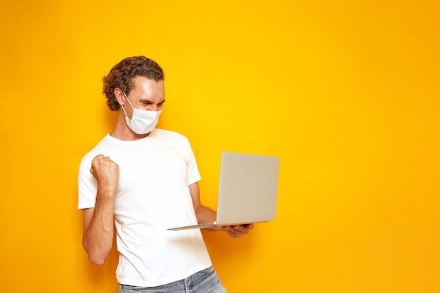 homme en masque médical vêtements décontractés avec ordinateur portable à la main se réjouit de la victoire sur fond jaune