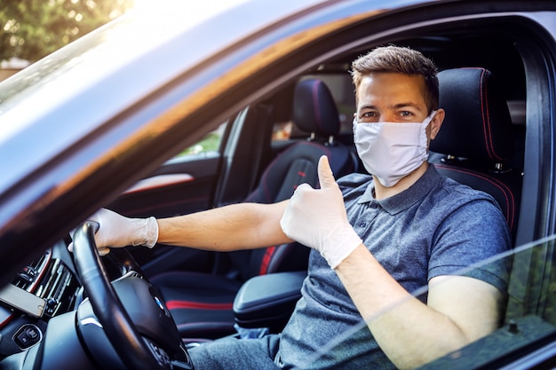 Homme avec masque de protection et gants au volant d'une voiture. épidémie. Restez prudent en affichant les pouces vers le haut.