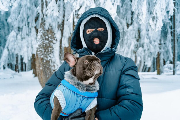 Photo homme avec un masque de ski et un bouledogue français dans la forêt d'hiver