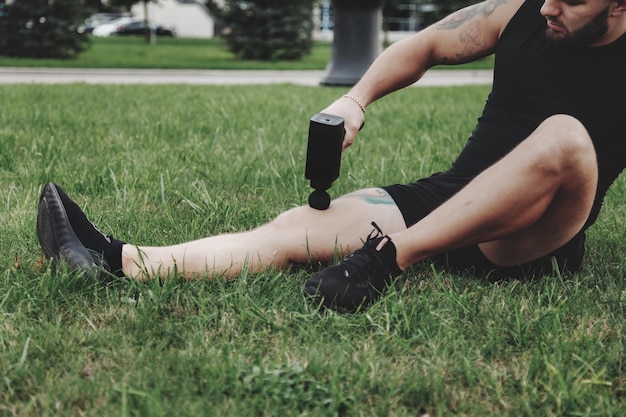 L'homme masse le corps avec un pistolet à percussion pendant les sports de plein air actifs. Pistolet de massage pour l'auto-récupération des performances musculaires pendant l'entraînement. Physiothérapie des athlètes. Activités de plein air