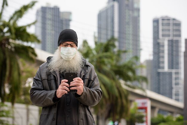 Homme mature hipster barbu avec masque à l'aide de téléphone à l'extérieur