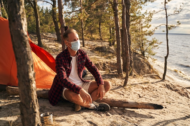 Un homme mature porte un masque médical alors qu'il est assis près d'une tente dans la forêt et regarde la mer devant