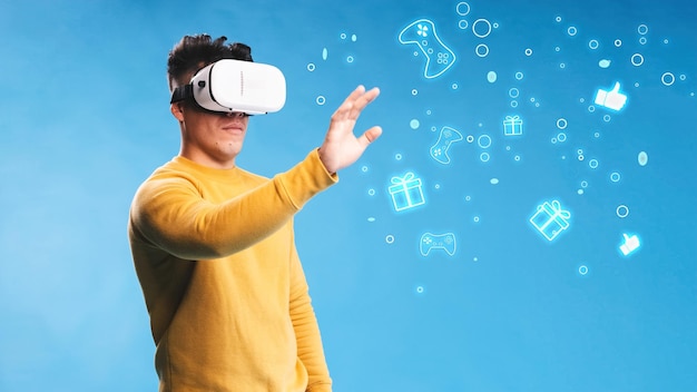 Homme méconnaissable dans des lunettes VR interagissant avec la réalité virtuelle contre des icônes de cadeaux avec des contrôleurs et comme bouton sur fond bleu