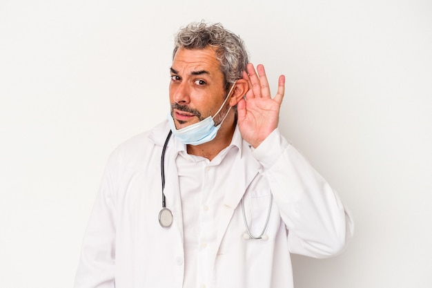 Homme médecin d'âge moyen portant un masque pour virus isolé sur fond blanc essayant d'écouter un potin.