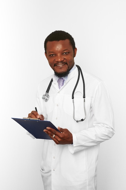 Homme médecin barbu noir souriant en robe blanche avec stéthoscope remplissant les dossiers médicaux sur le presse-papiers