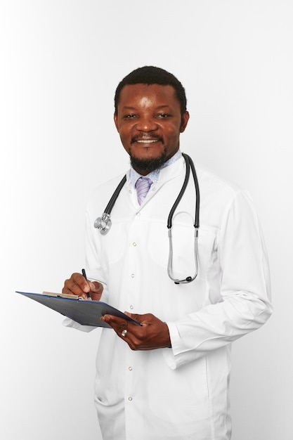 Photo homme médecin barbu noir souriant en robe blanche avec stéthoscope remplissant les dossiers médicaux sur le presse-papiers